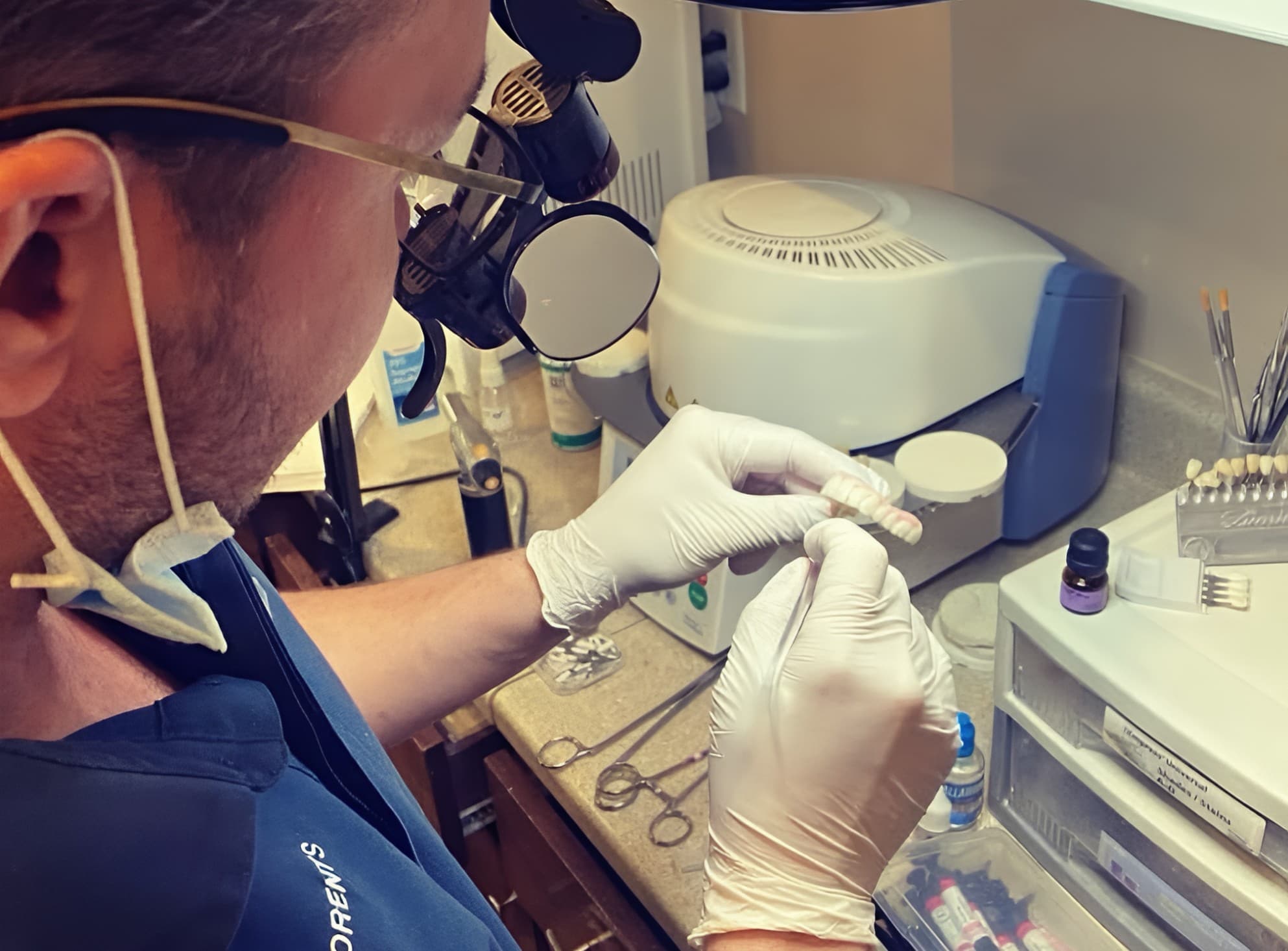 Dr. Edward Lorents, wearing loupes, checking a finished arch against a shade guide at the bench beside the ceramic furnace